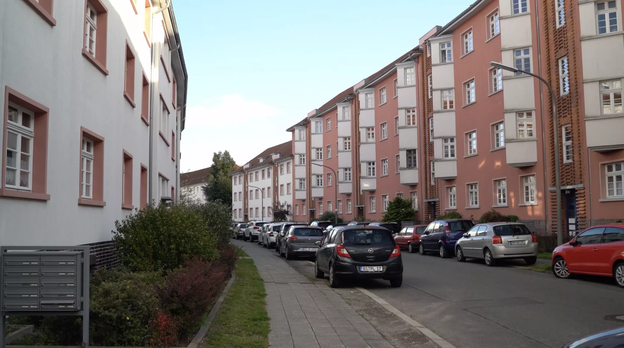 Eine Wohnstraße mit geparkten Autos, rosafarbenen und weißen Wohnhäusern und einem von Büschen gesäumten Bürgersteig unter einem klaren Himmel.