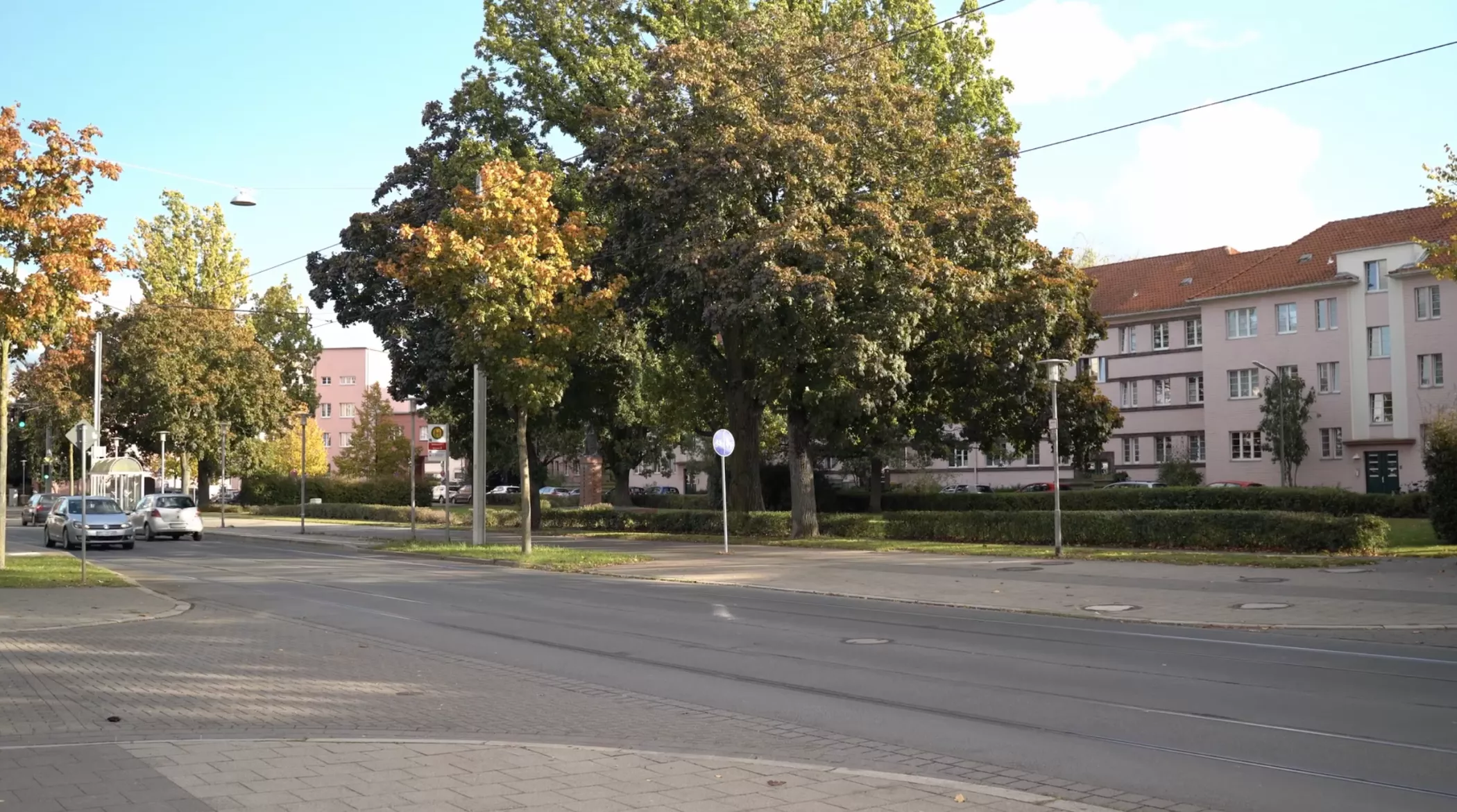 Eine ruhige städtische Straße mit Straßenbahnschienen, grünen und herbstlich belaubten Bäumen und Wohngebäuden im Hintergrund.