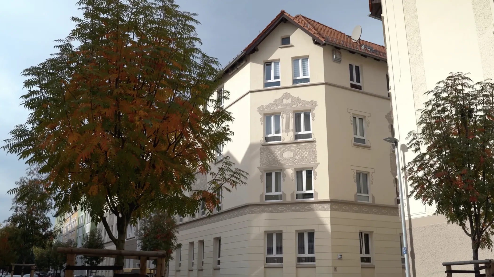 Ein cremefarbenes, dreistöckiges Wohnhaus mit rotem Ziegeldach, neben einem Baum mit grünen und orangefarbenen Blättern.