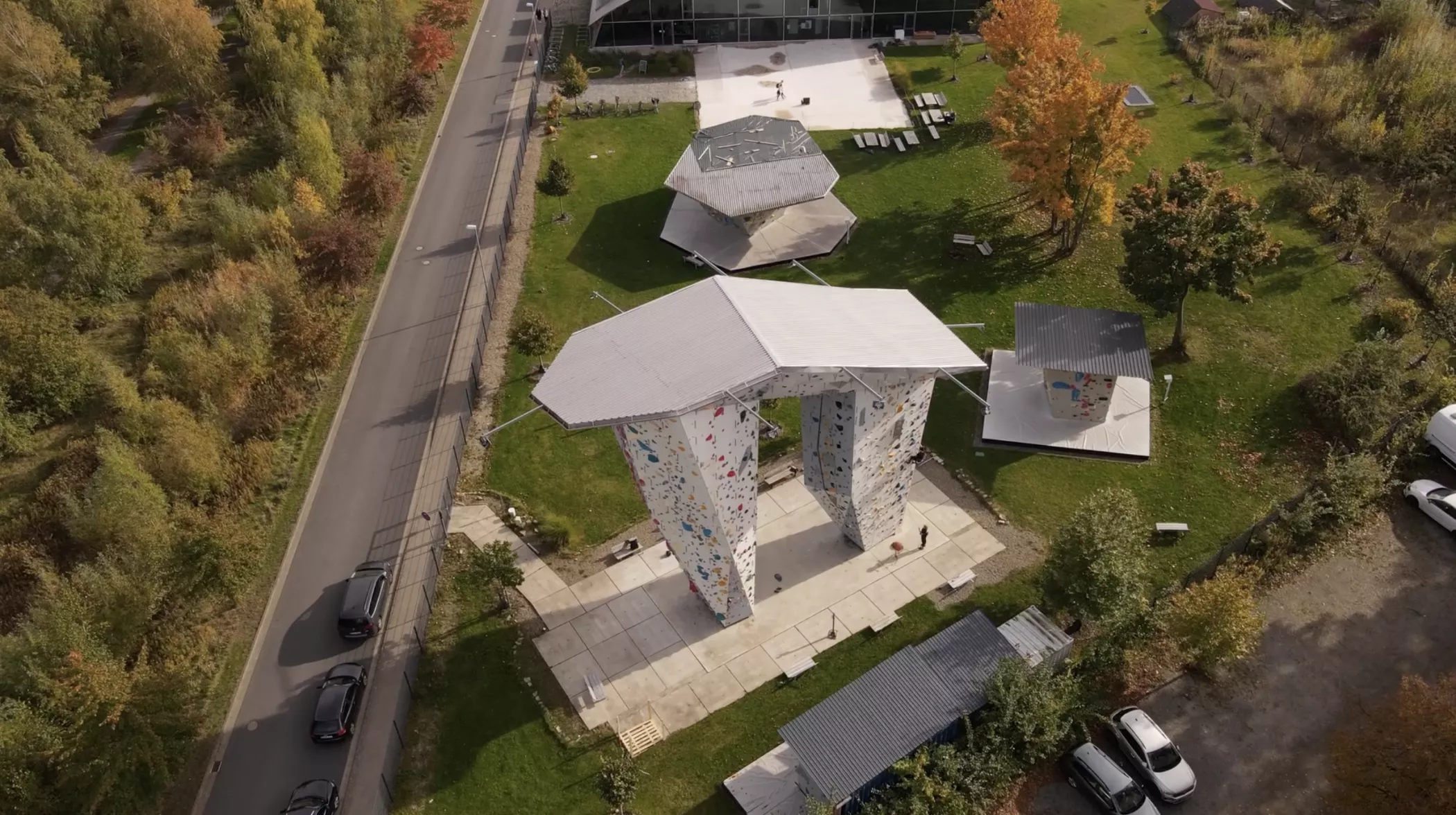 Aerial view of an outdoor climbing wall with geometric roofs and adjacent hexagonal shelters, surrounded by grass, trees and a road.