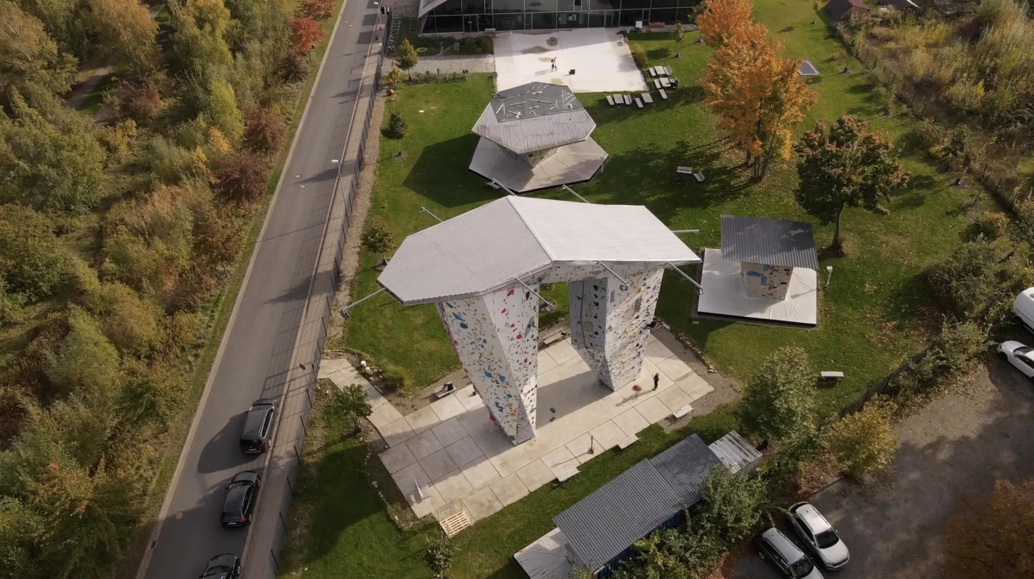 Luftaufnahme einer Outdoor-Kletterwand mit geometrischen Dächern und angrenzenden sechseckigen Unterständen, umgeben von Gras, Bäumen und einer Straße.
