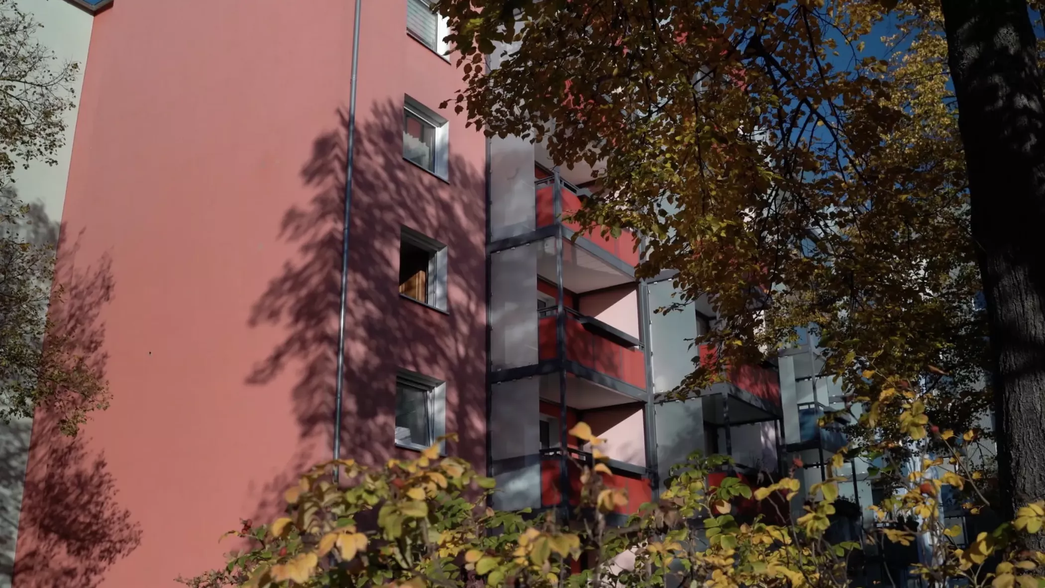 Açık mavi bir gökyüzü altında, yaprak döken ağaçların kısmen gölgelediği balkonlara sahip pembe bir konut binası.