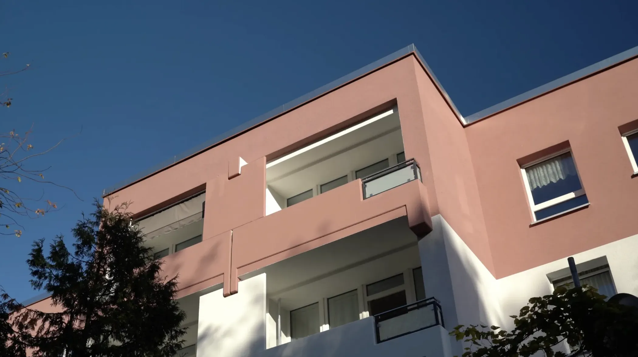 Ein modernes, rosa-weißes Wohnhaus mit Balkonen, von denen aus man den blauen Himmel überblicken kann.
