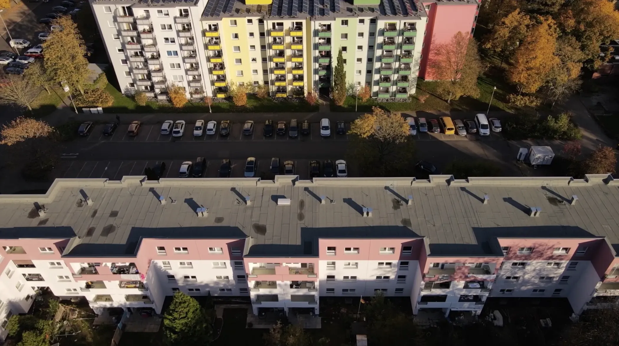 Eine Luftaufnahme von Wohngebäuden mit geparkten Autos und Bäumen dazwischen, aufgenommen an einem sonnigen Tag.
