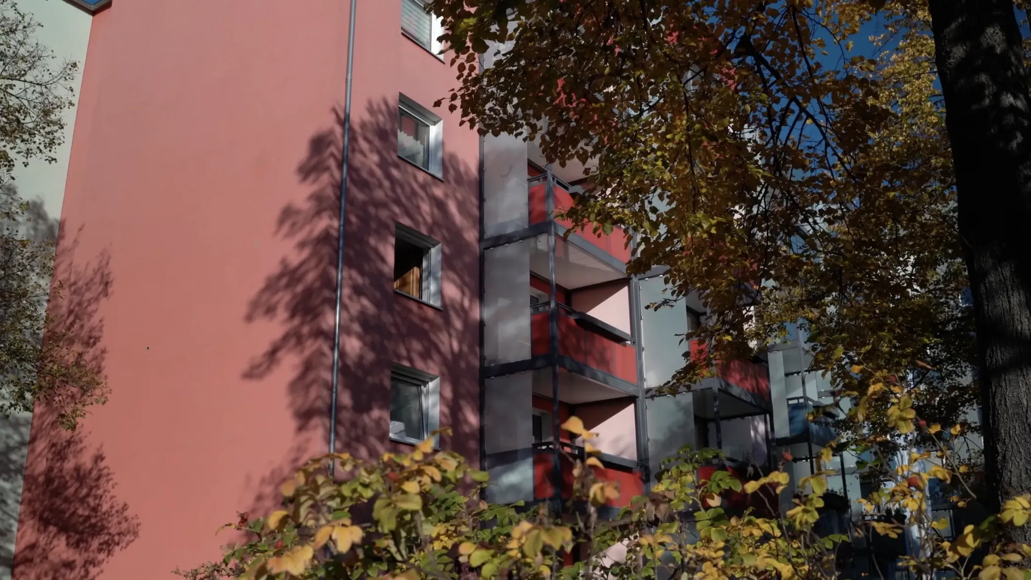 Ein rosafarbenes Wohnhaus mit Balkonen, die teilweise von Laubbäumen beschattet werden, unter einem klaren blauen Himmel.