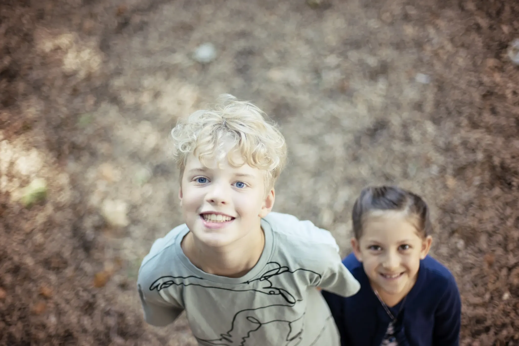 Two smiling children look into the camera outdoors. They are standing on a floor covered with brown leaves or wood shavings.