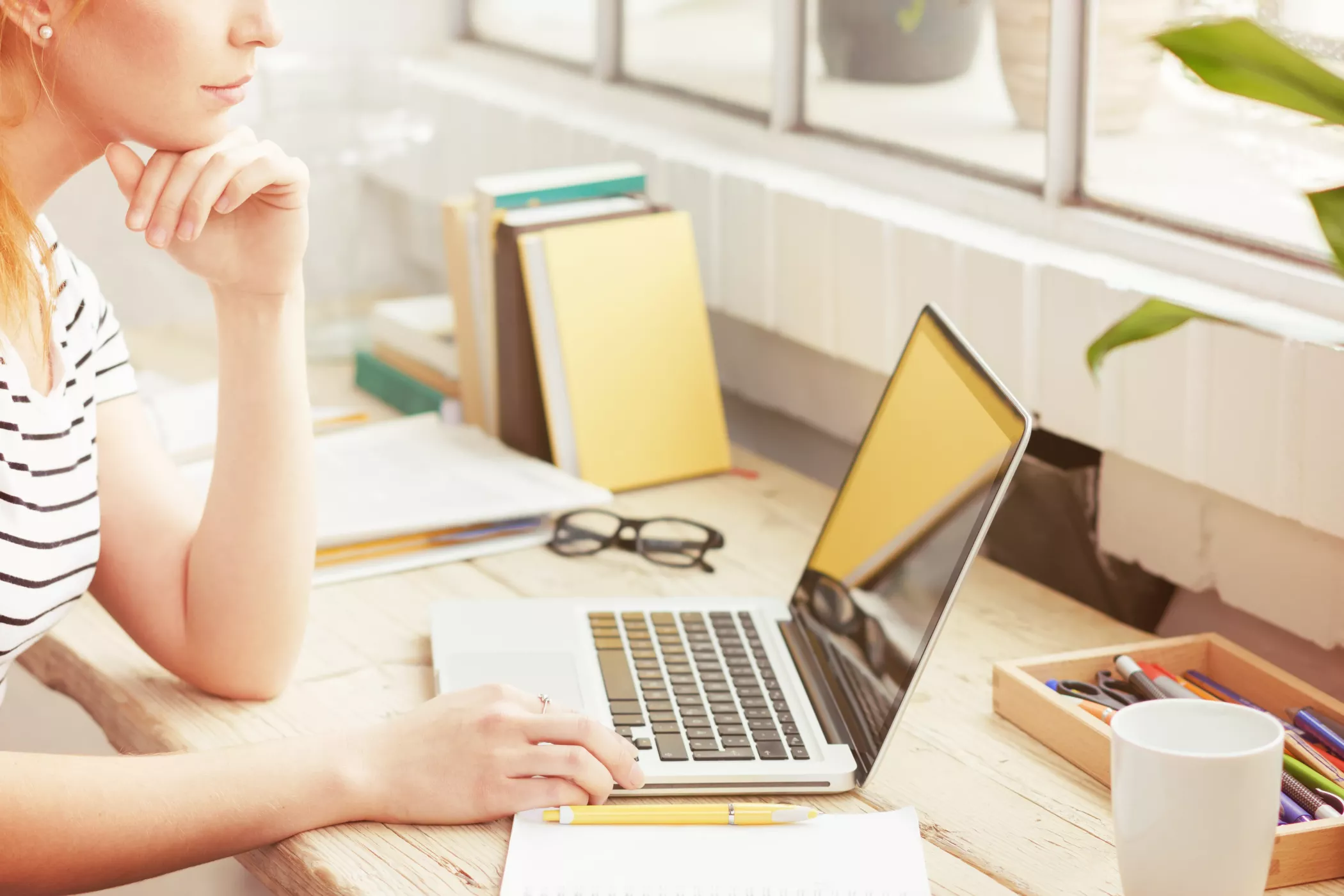 Eine Frau sitzt an einem hölzernen Schreibtisch mit einem Laptop, Büchern und Schreibwaren und arbeitet konzentriert vor einem sonnendurchfluteten Fenster.