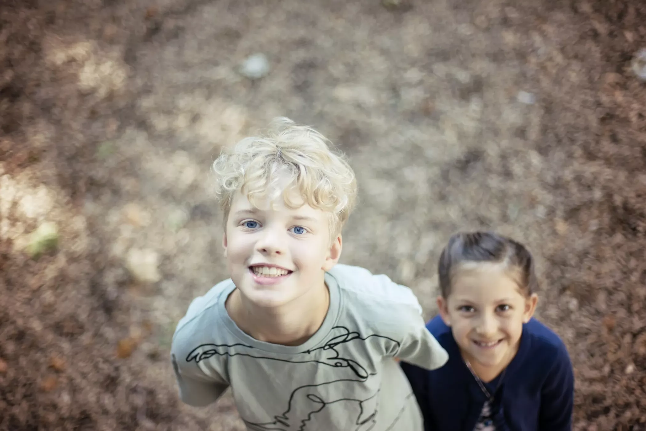 Deux enfants souriants regardent la caméra en extérieur. Ils sont debout sur un sol recouvert de feuilles brunes ou de copeaux de bois.