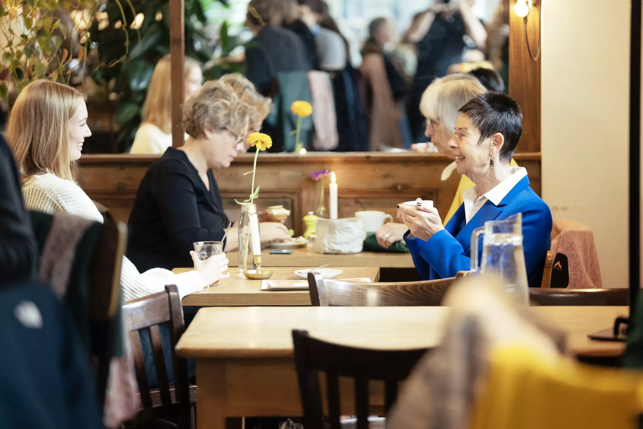 Drei Frauen sitzen lächelnd und plaudernd an einem Cafétisch, mit Getränken und Blumen auf dem Tisch in einem überfüllten, warm beleuchteten Raum.