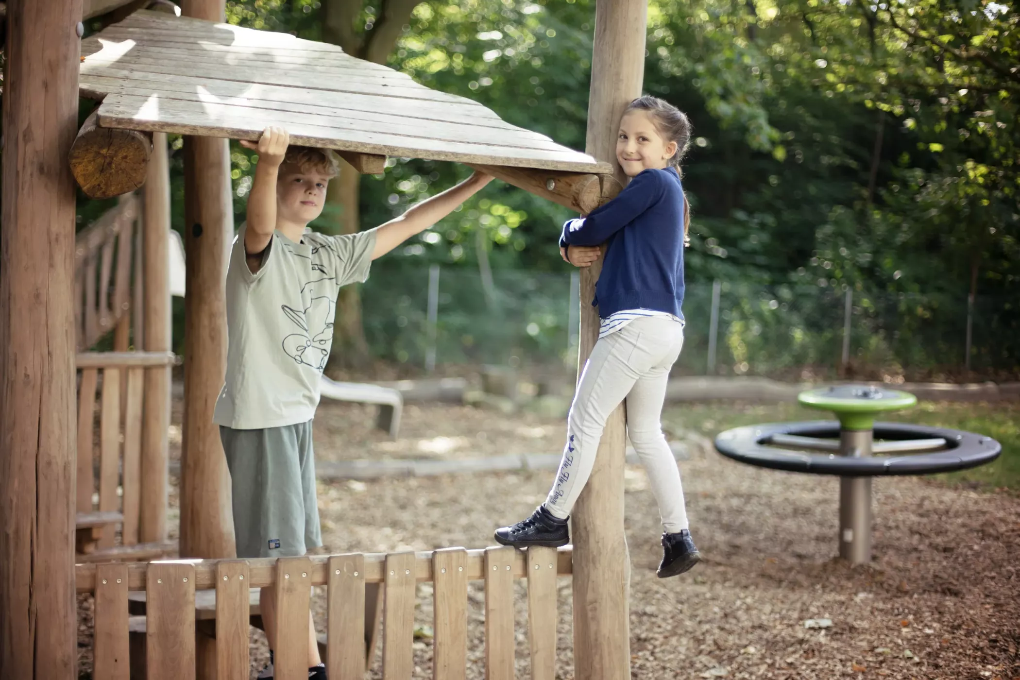 Zwei Kinder spielen auf einem hölzernen Spielplatz; eines hängt von der Decke, das andere klammert sich an eine Stange, beide lächeln.