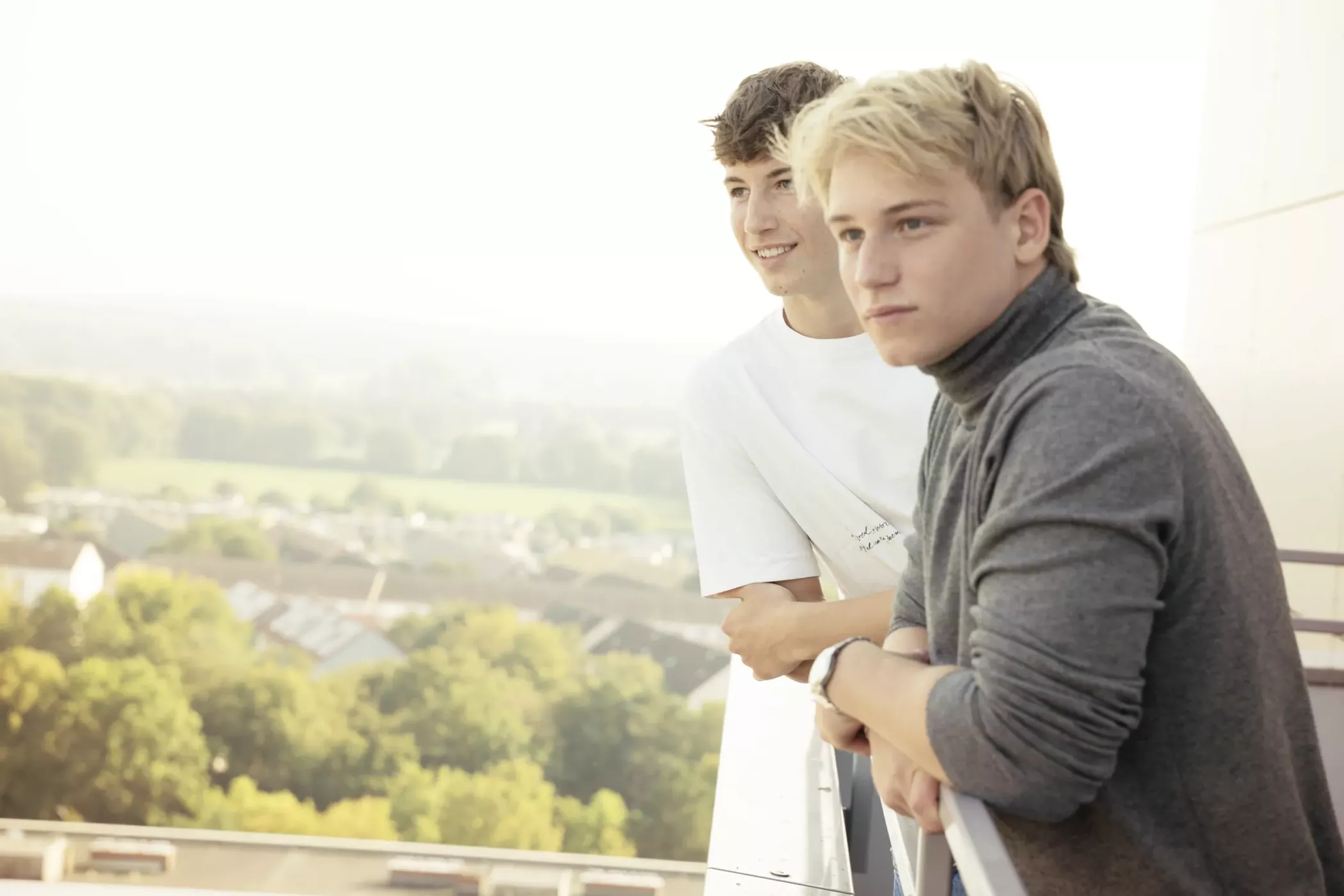 Deux jeunes hommes se tiennent sur un balcon, s'appuient sur la balustrade et regardent attentivement le paysage en dessous d'eux.