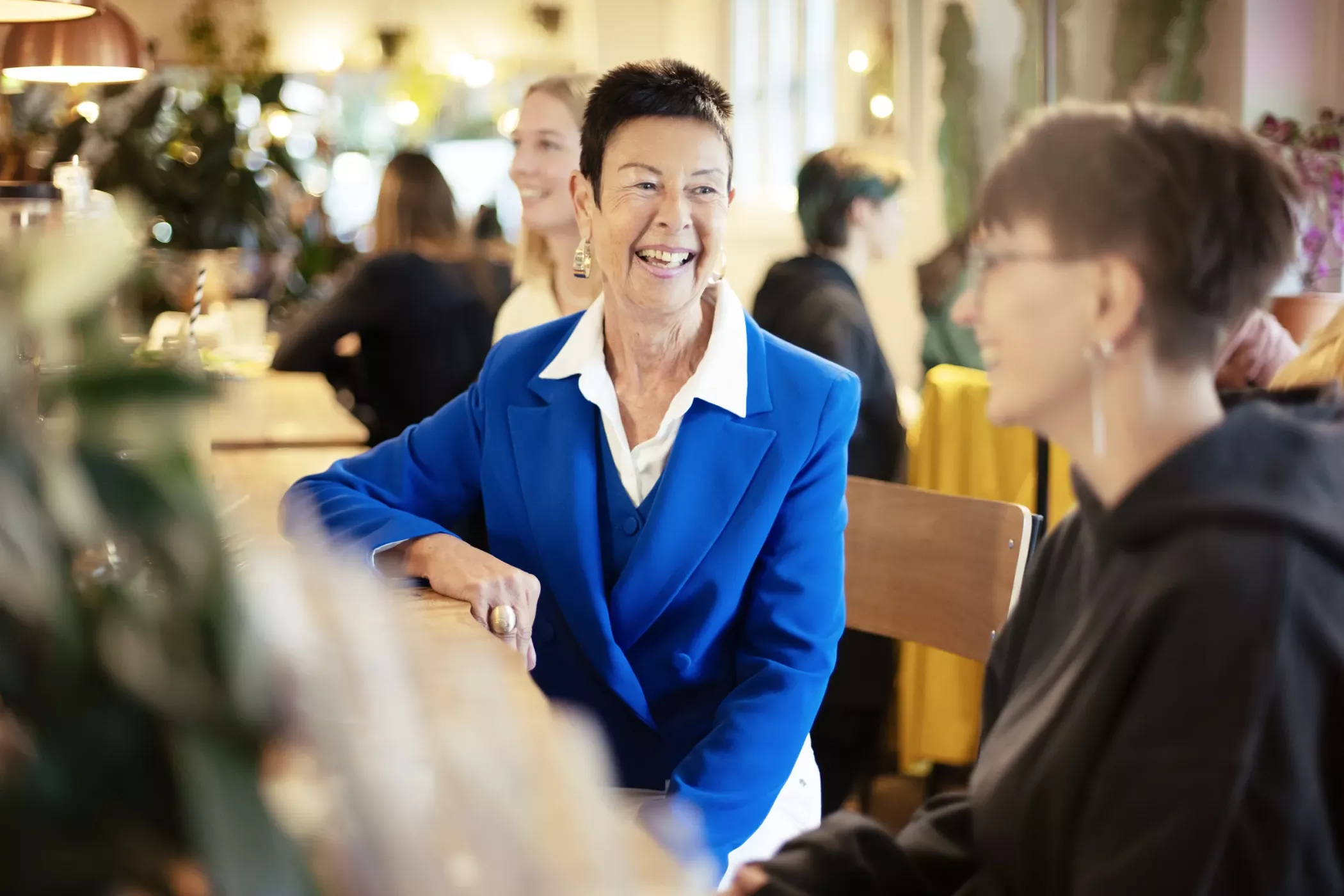 Une femme d'un certain âge, vêtue d'un tailleur bleu, sourit en discutant avec une amie dans un café très fréquenté.
