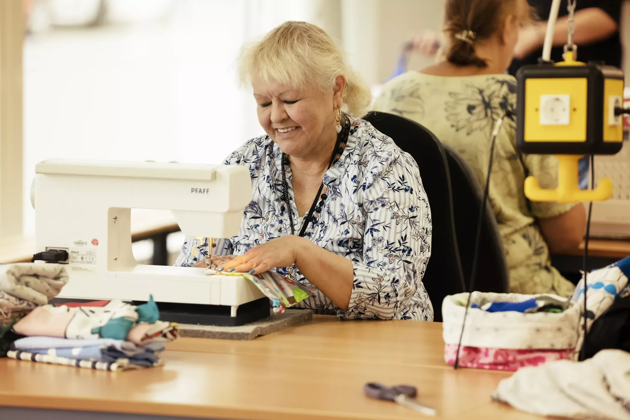 Une femme sourit en cousant du tissu avec une machine à coudre posée sur une table, entourée de tissus pliés et d'outils de couture.