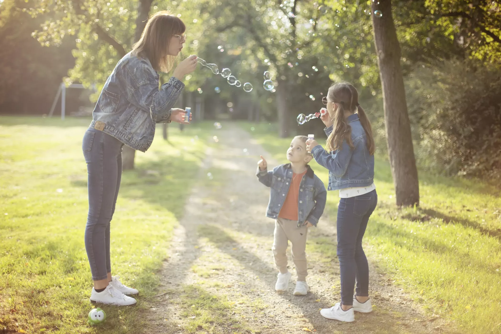 Une femme et deux enfants font des bulles ensemble sur un chemin de jardin ensoleillé, tous vêtus de jeans et de vêtements décontractés.