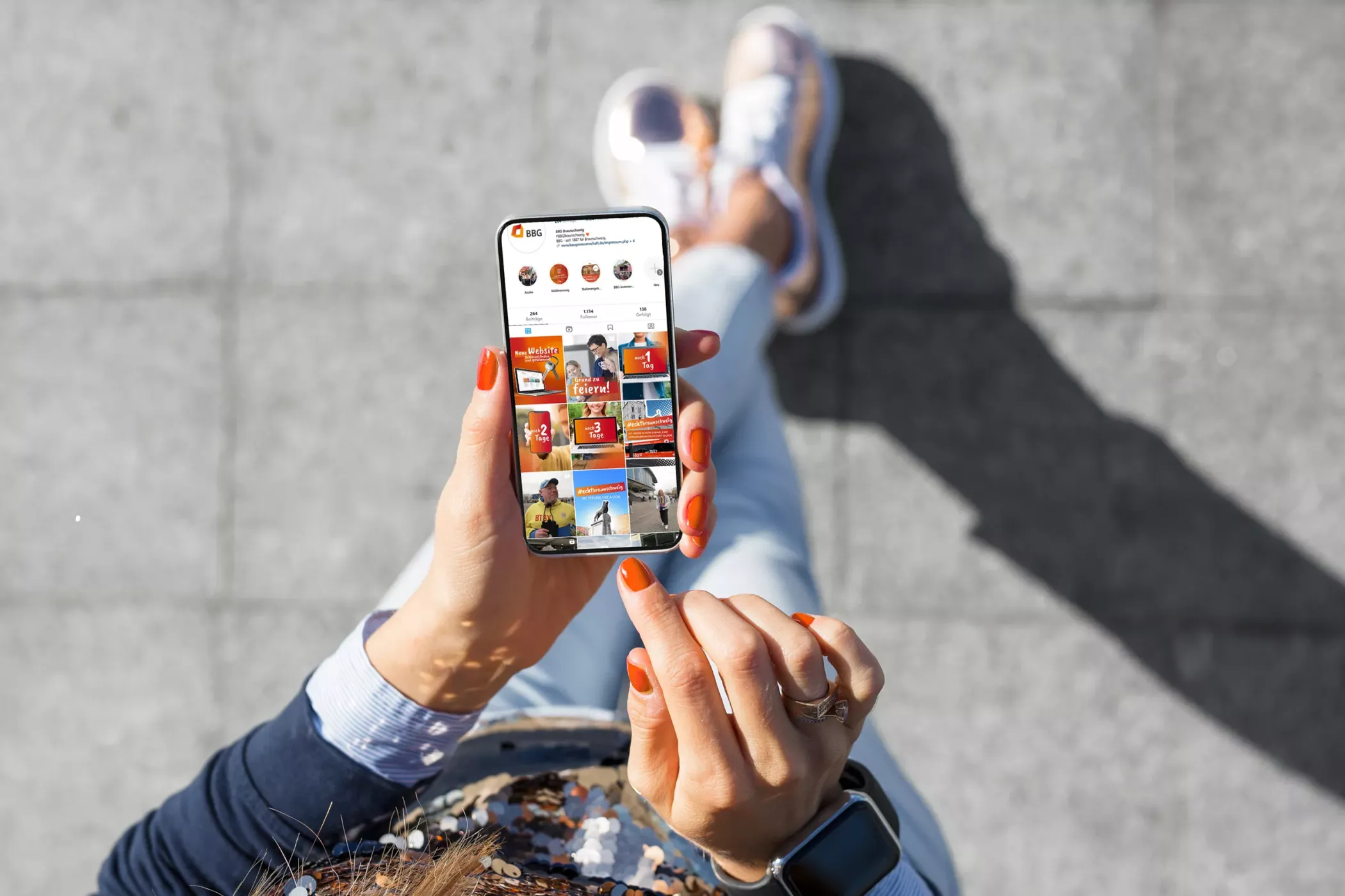 Eine Person mit orange lackierten Nägeln hält ein Smartphone in der Hand und scrollt durch ein Instagram-Konto, während sie draußen auf einem grauen Bürgersteig sitzt.