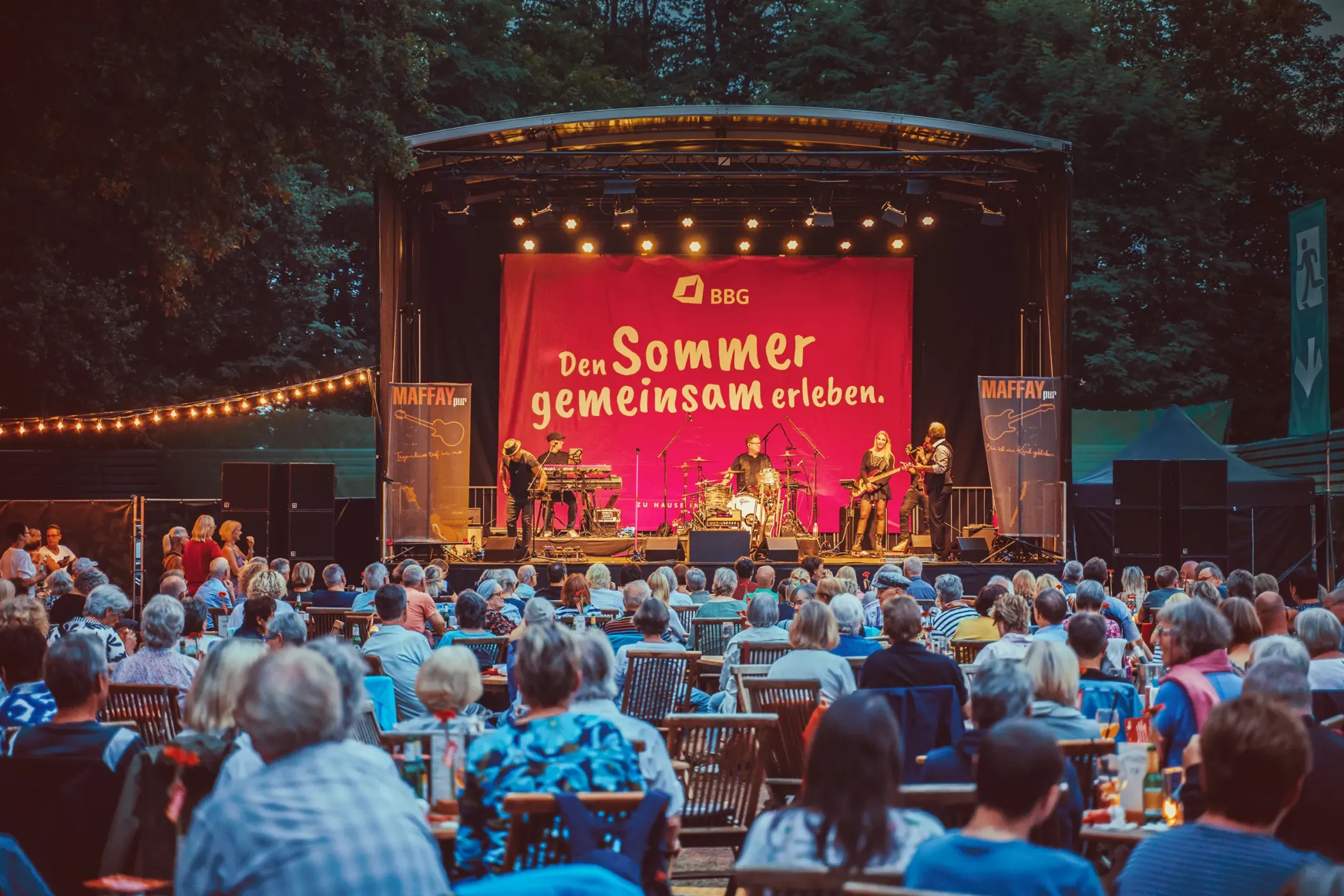 Eine Menschenmenge beobachtet einen Live-Auftritt einer Band auf einer Open-Air-Bühne mit der Aufschrift 