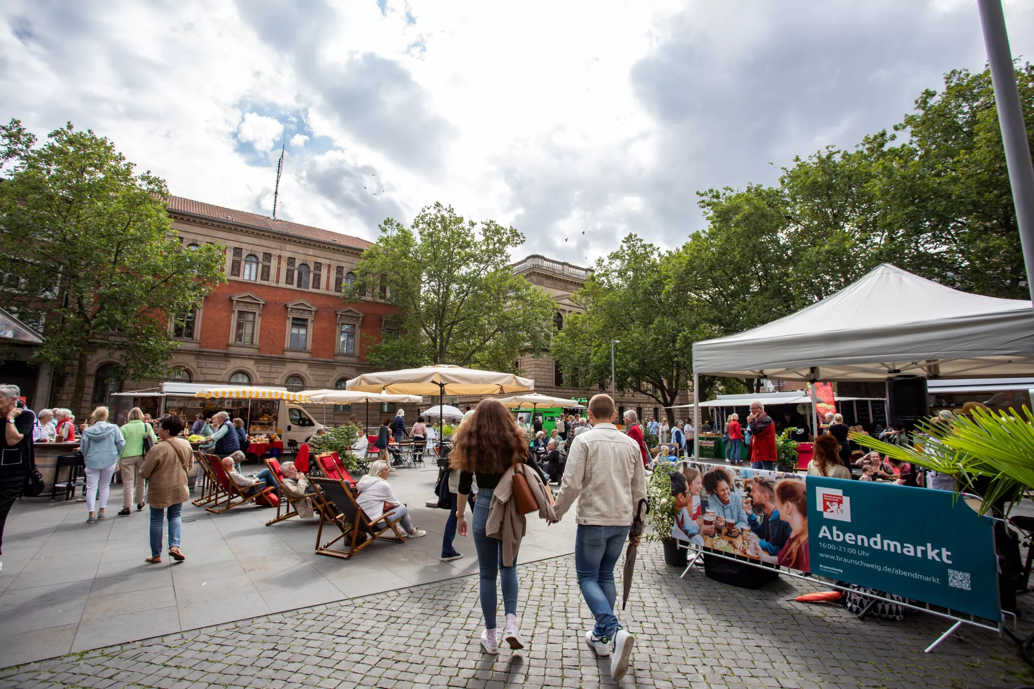 Unter teilweise bewölktem Himmel, umgeben von historischen Gebäuden und Bäumen, genießen die Menschen einen Markt unter freiem Himmel mit Essensständen, Bänken und Zelten.