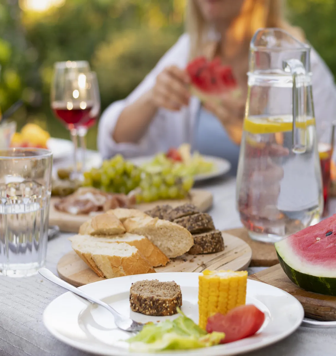 Ein im Freien gedeckter Tisch mit Brot, Mais, Obst, Zitronenwasser und einer Person, die eine Scheibe Wassermelone in der Hand hält, im Hintergrund.