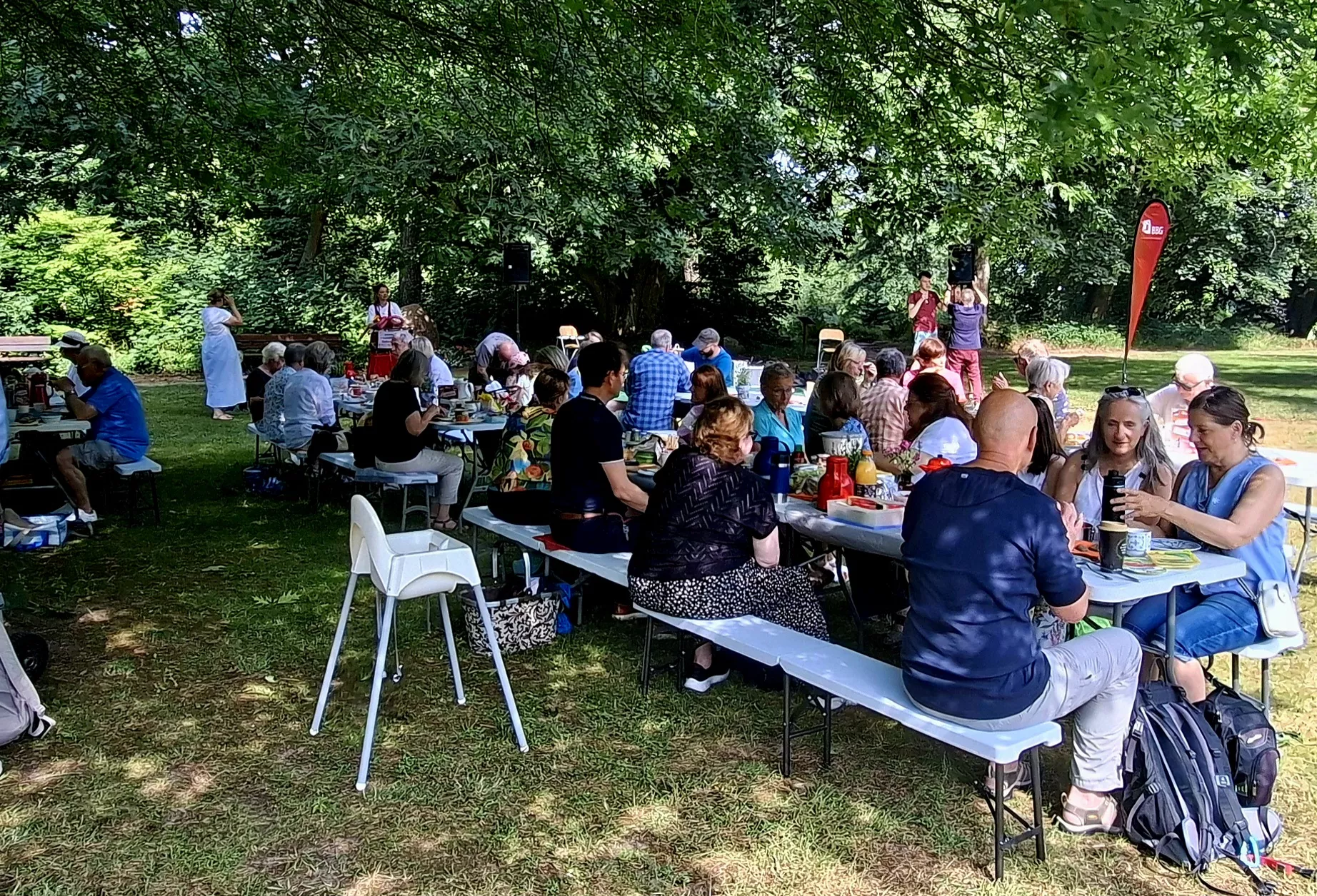 Eine Gruppe von Menschen sitzt an Picknicktischen unter Bäumen, isst und unterhält sich bei einem Treffen im Freien in einem Park.