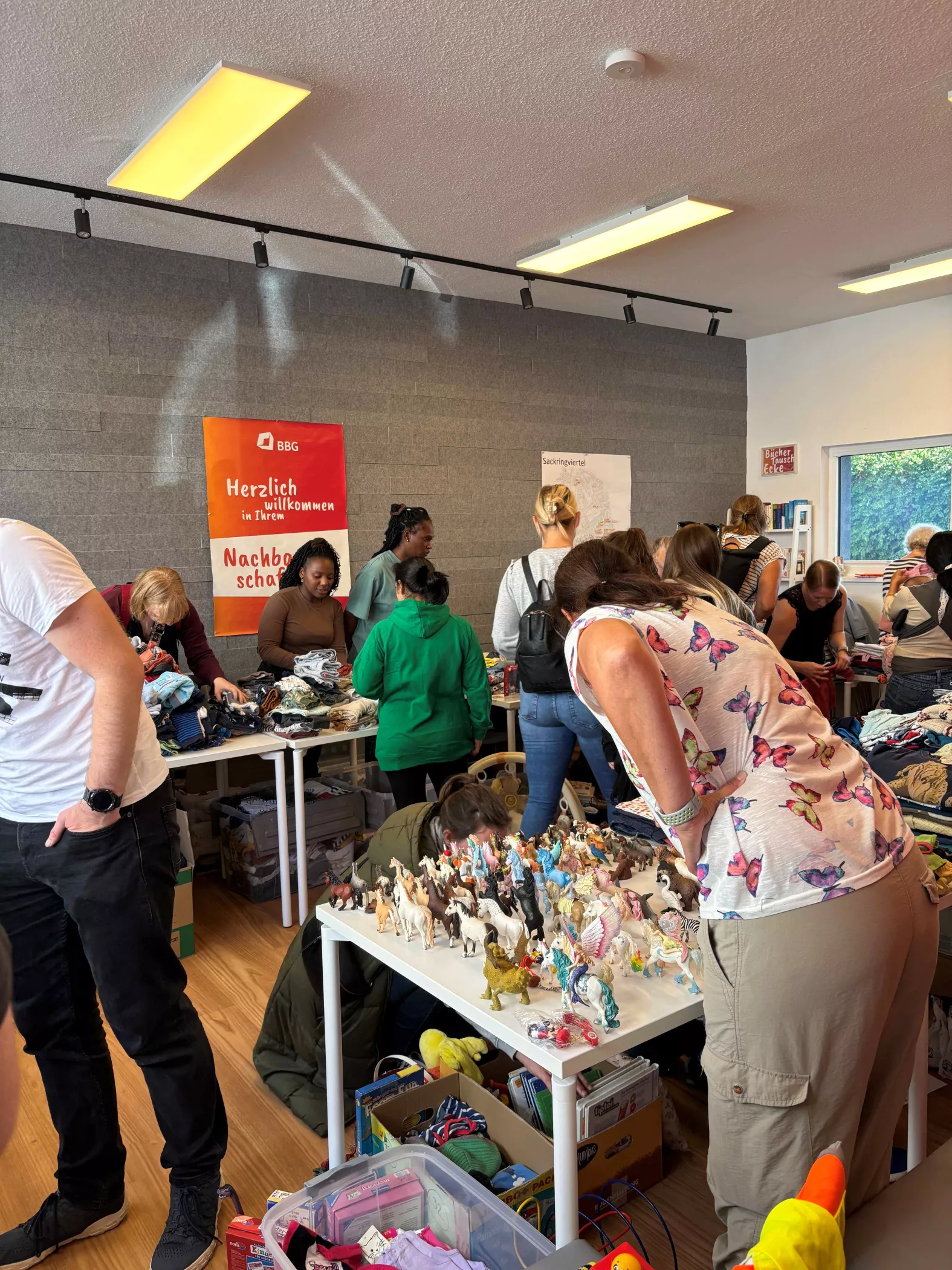 Die Leute stöbern auf einem Indoor-Flohmarkt durch Tische mit Spielzeug und Kleidung; ein rotes Willkommensschild hängt an der Wand.