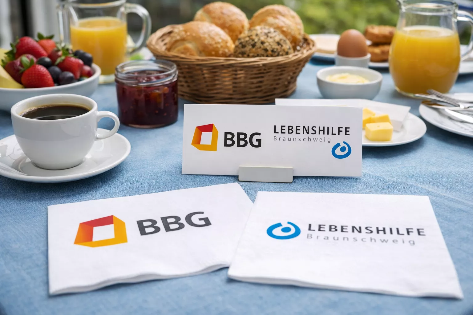 Ein Frühstückstisch mit Kaffee, Saft, Obst, Gebäck und Servietten mit den Logos der BBG und der Lebenshilfe Braunschweig.