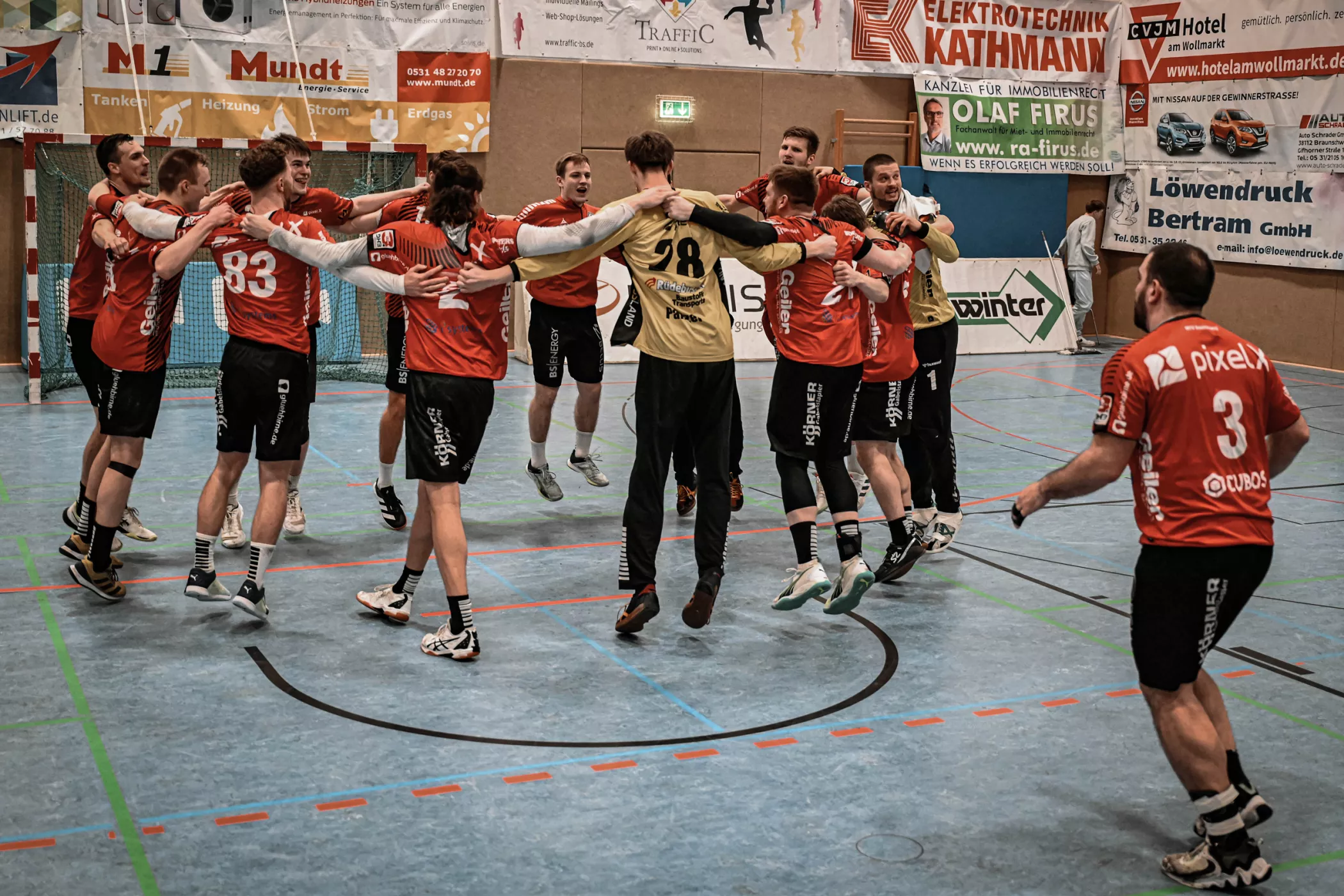 Eine Männerhandballmannschaft in roten Trikots feiert im Kreis, springt und hält sich an den Händen auf einem Hallenplatz.