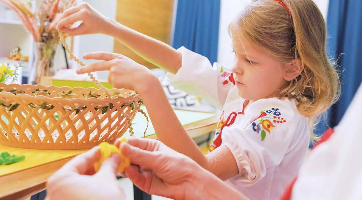 Ein kleines Mädchen in bestickten Kleidern sitzt an einem Tisch und hält Perlen in der Nähe eines Weidenkorbs, unterstützt von den Händen eines Erwachsenen.