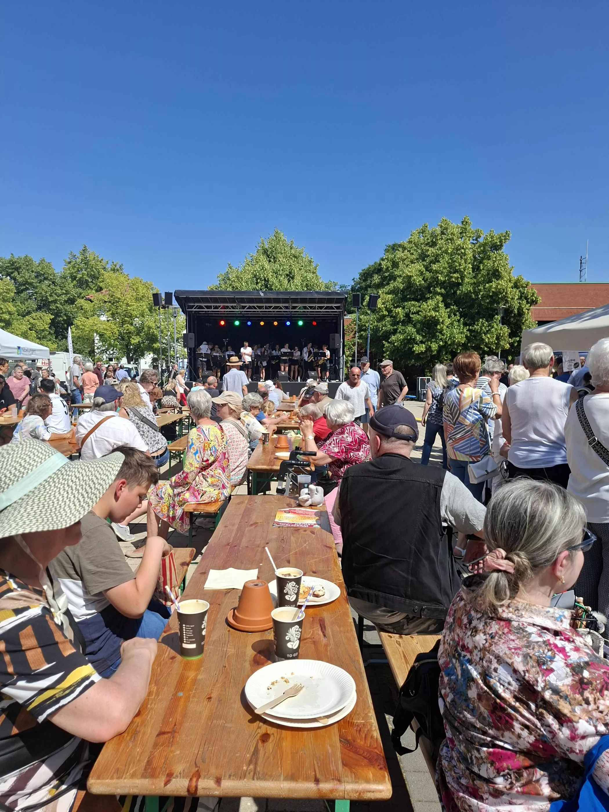 Die Menschen sitzen an Tischen im Freien, essen und trinken und schauen einer Live-Band zu, die unter blauem Himmel auf der Bühne spielt.