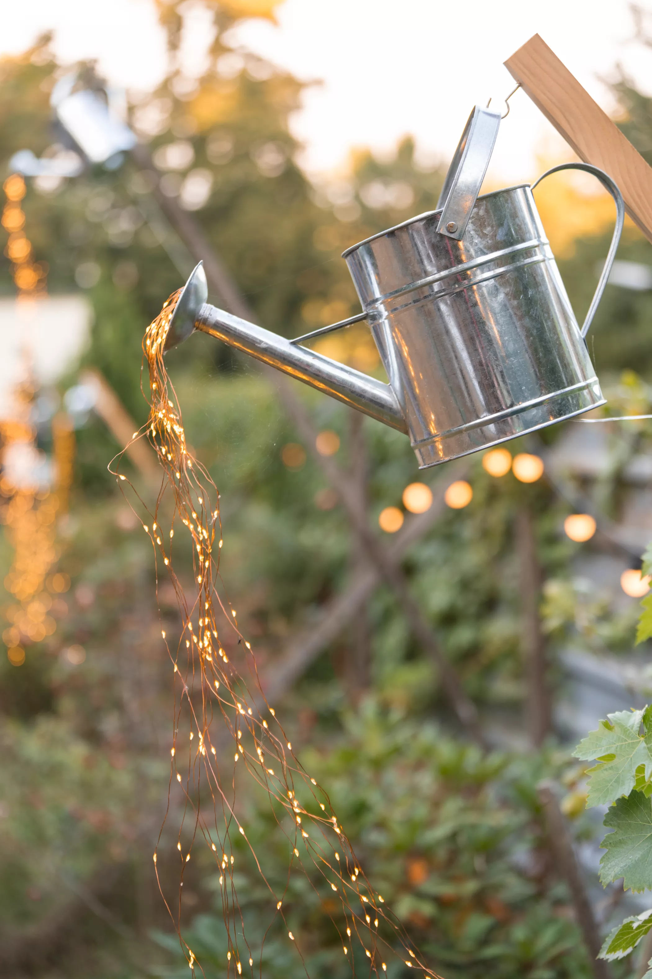 Eine Gießkanne aus Metall hängt im Freien, aus deren Ausguss eine Lichterkette als skurrile Gartendekoration herausragt.