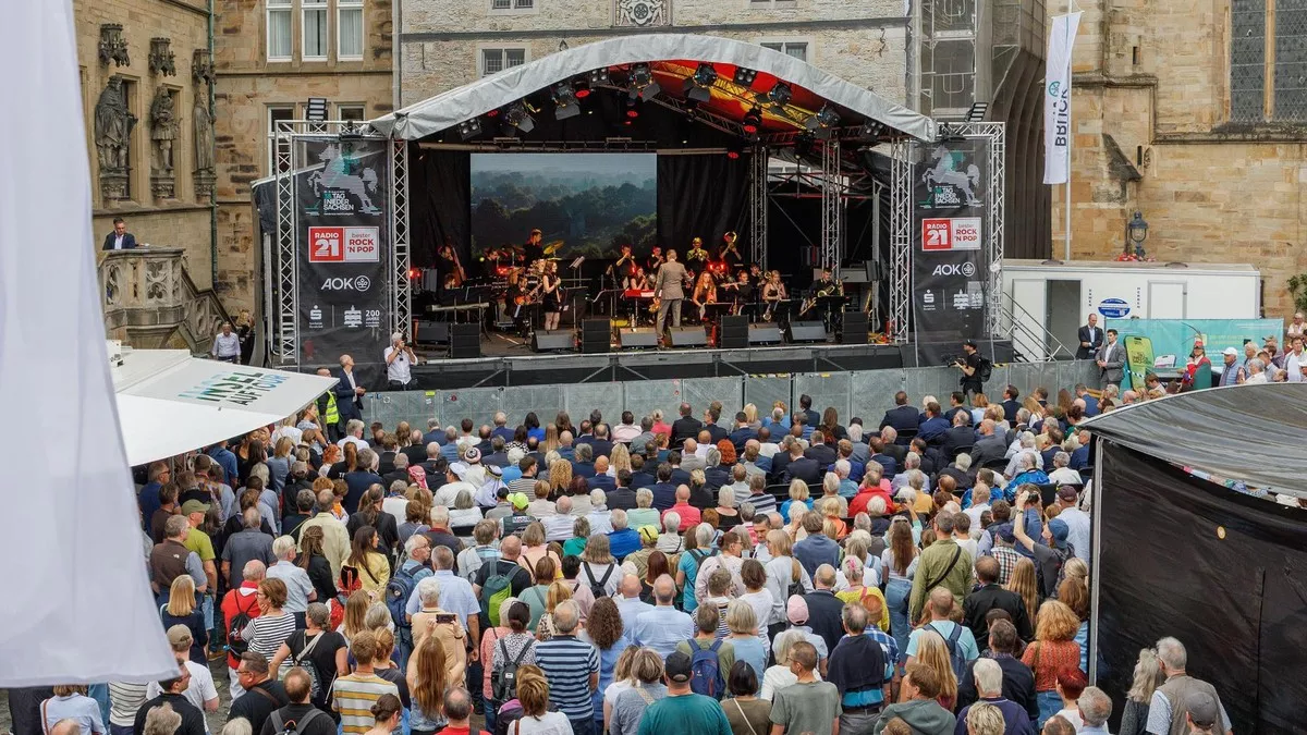 Eine große Menschenmenge verfolgt den Auftritt einer Live-Band auf einer Open-Air-Bühne bei einem Festival vor historischen Gebäuden.