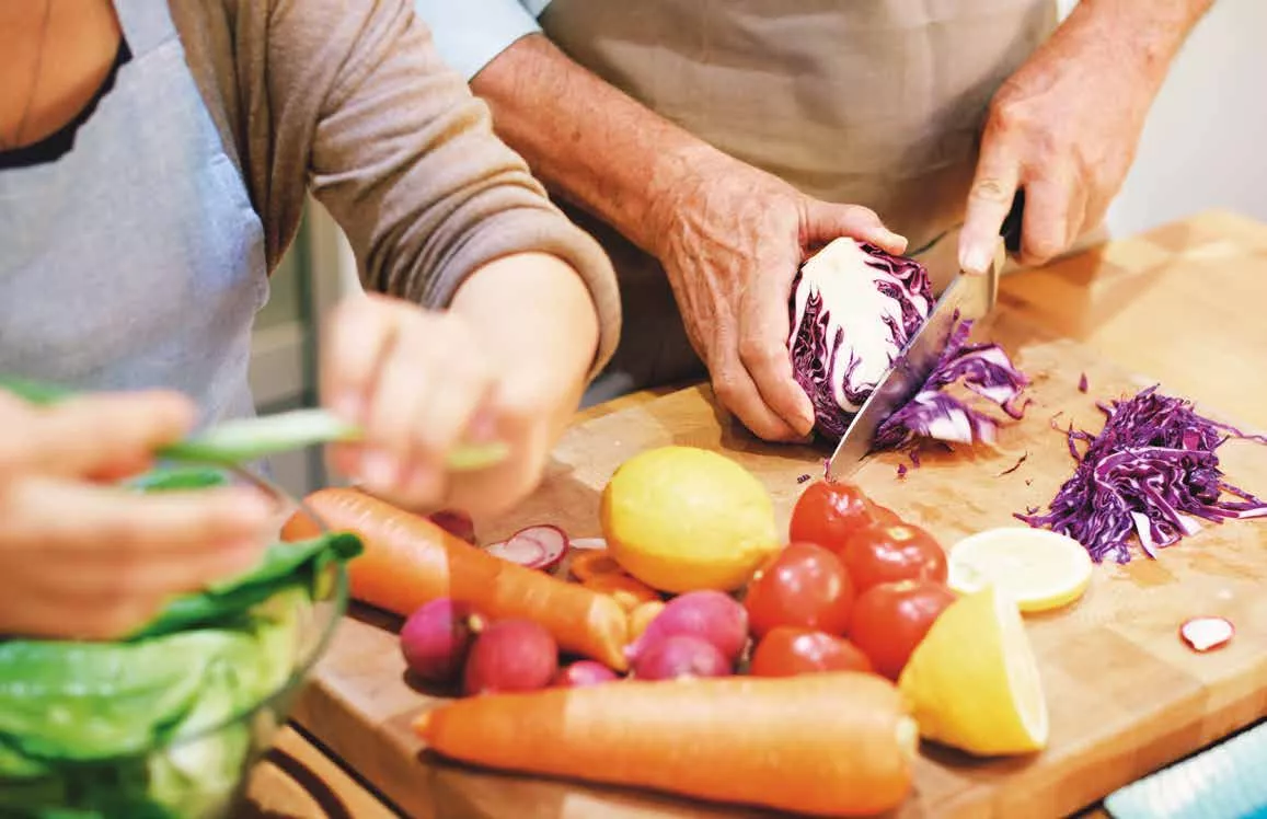 Zwei Personen schneiden frisches Gemüse, darunter Kohl, Karotten, Tomaten und Zitronen, auf einem hölzernen Schneidebrett.