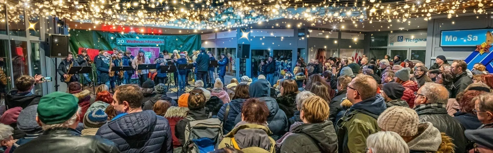 Eine große Menschenmenge versammelt sich unter Lichterketten, um den Musikern zuzusehen, die auf einer kleinen Bühne auf einem überdachten Weihnachtsmarkt auftreten.