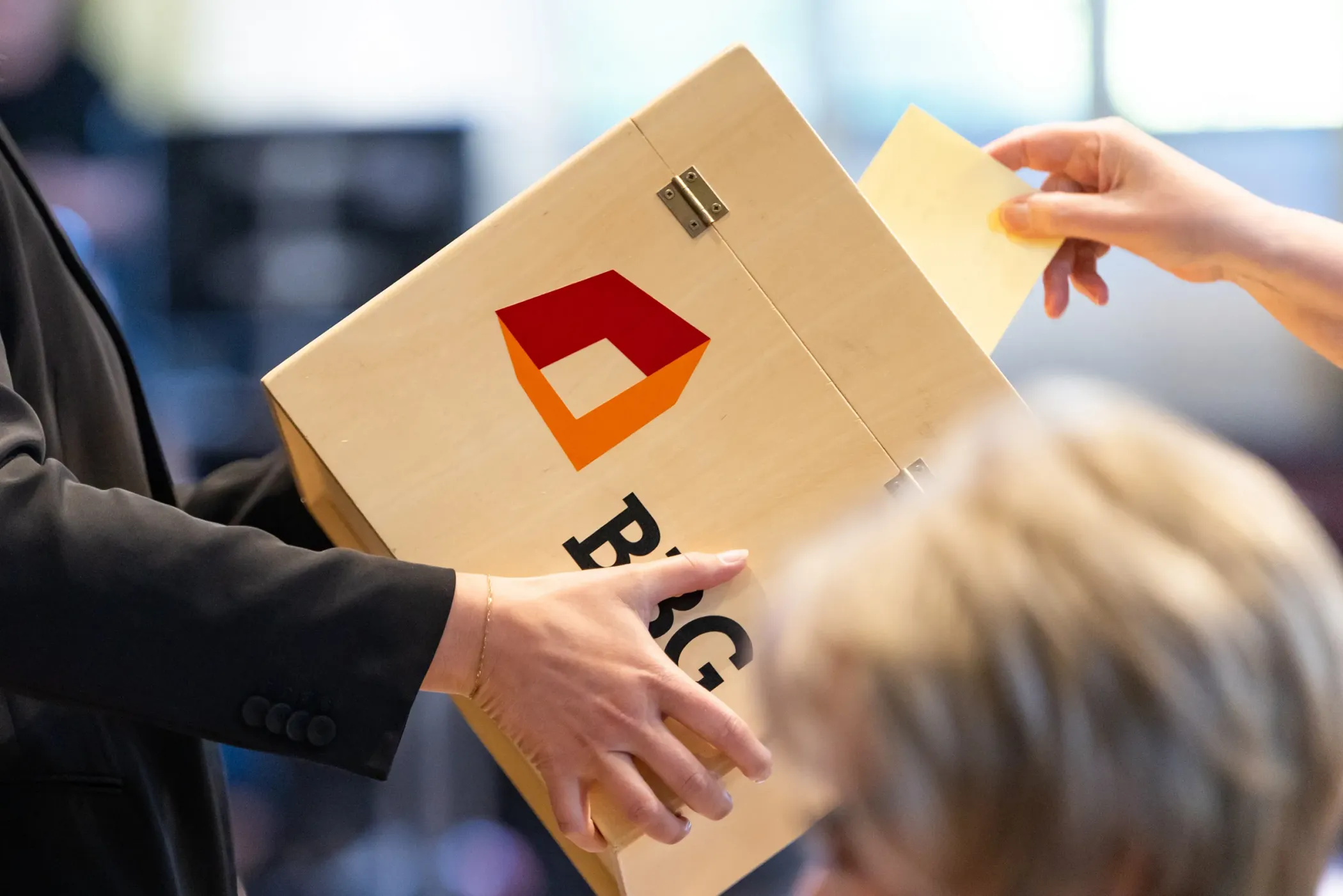 Eine Person wirft bei einer scheinbaren Wahl oder Abstimmung einen Stimmzettel in eine Holzkiste mit dem rot-orangenen BBG-Logo.
