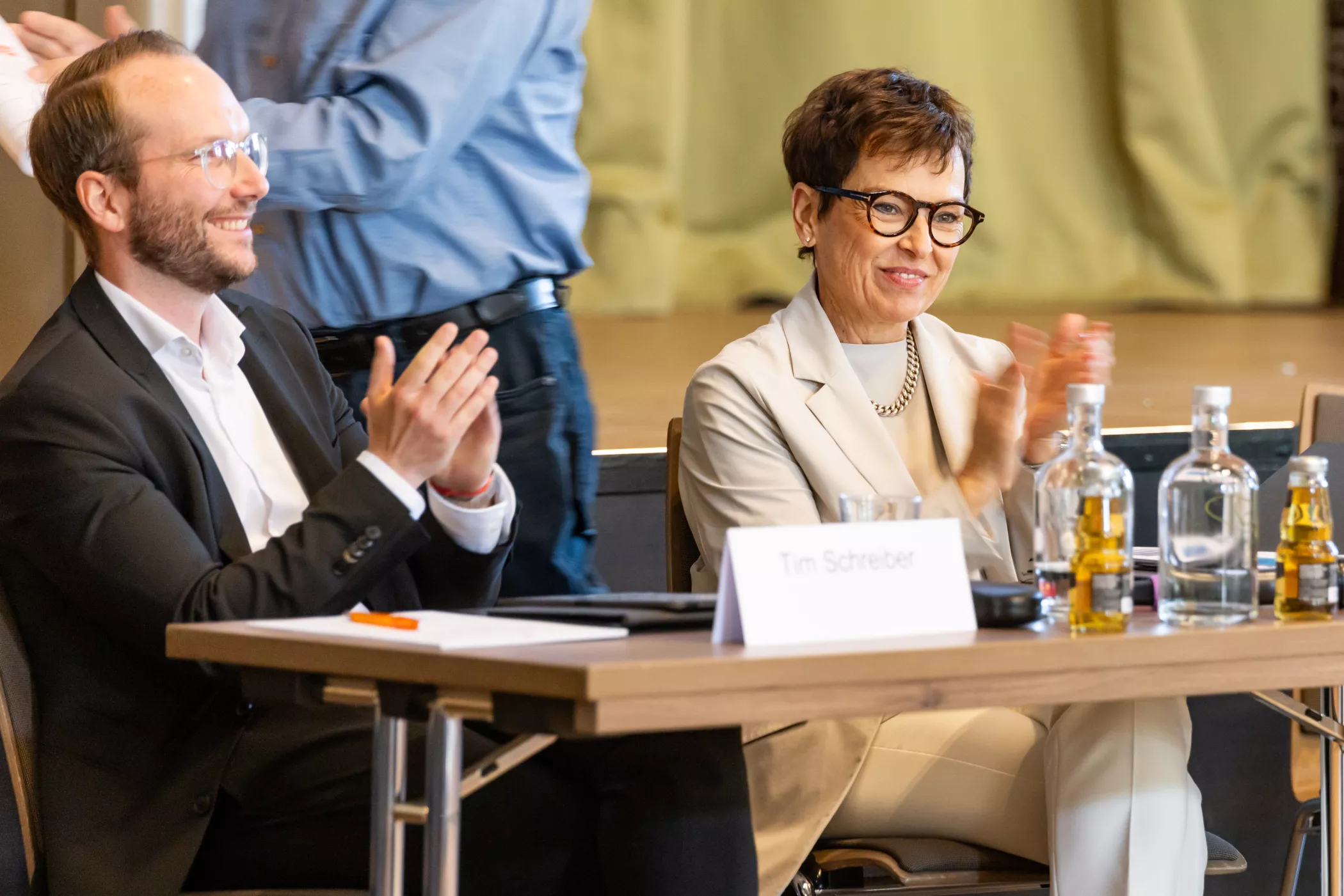 Zwei Personen in Geschäftskleidung sitzen an einem Tisch, lächeln und klatschen während einer Veranstaltung. Flaschen und Namensschild auf dem Tisch.