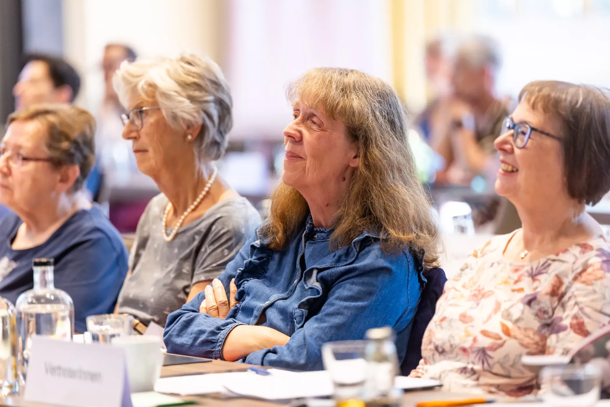 Vier ältere Frauen sitzen an einem Tisch, lächeln und hören während einer Sitzung oder eines Seminars in einem hellen Raum aufmerksam zu.