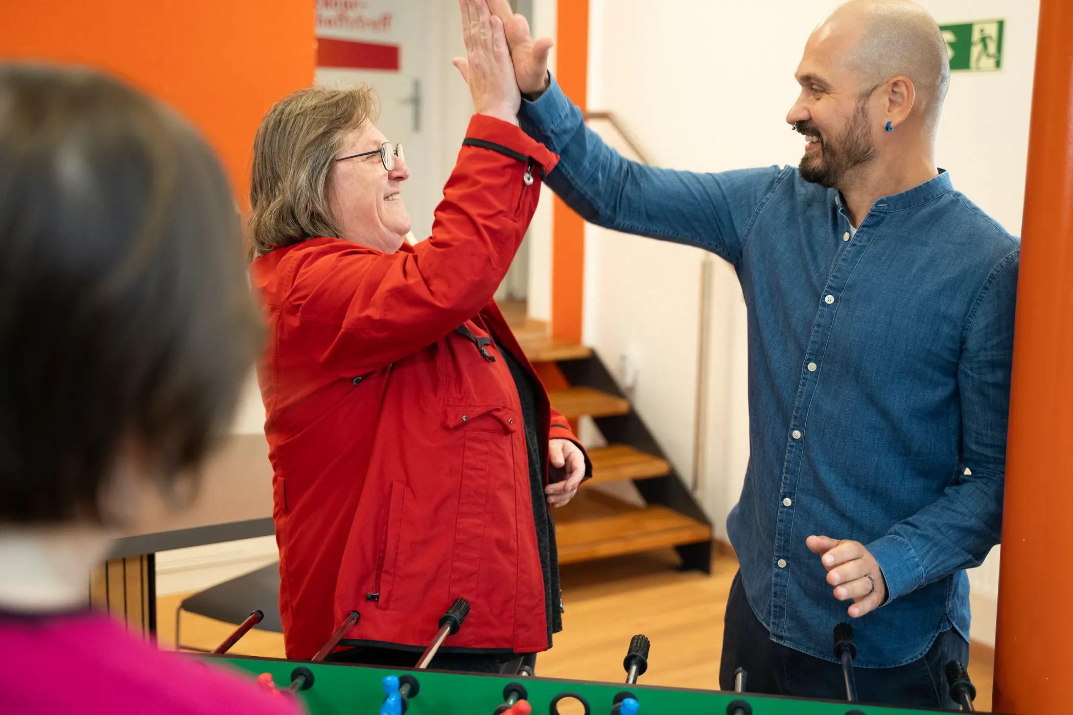 Zwei Menschen lächeln und schütteln sich die Hände neben einem Tischfußballtisch in einem hell erleuchteten Raum.