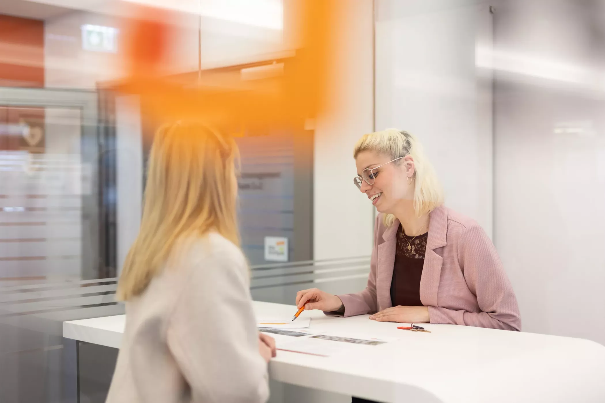 Zwei Frauen sitzen an einem modernen Schreibtisch in einem Büro, eine lächelt und hält einen Bleistift in der Hand, während die andere Papierkram bespricht.
