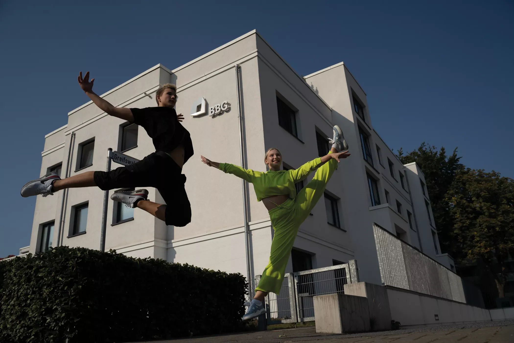 Two people in sportswear jump and pose energetically in front of a modern white building under a clear blue sky.
