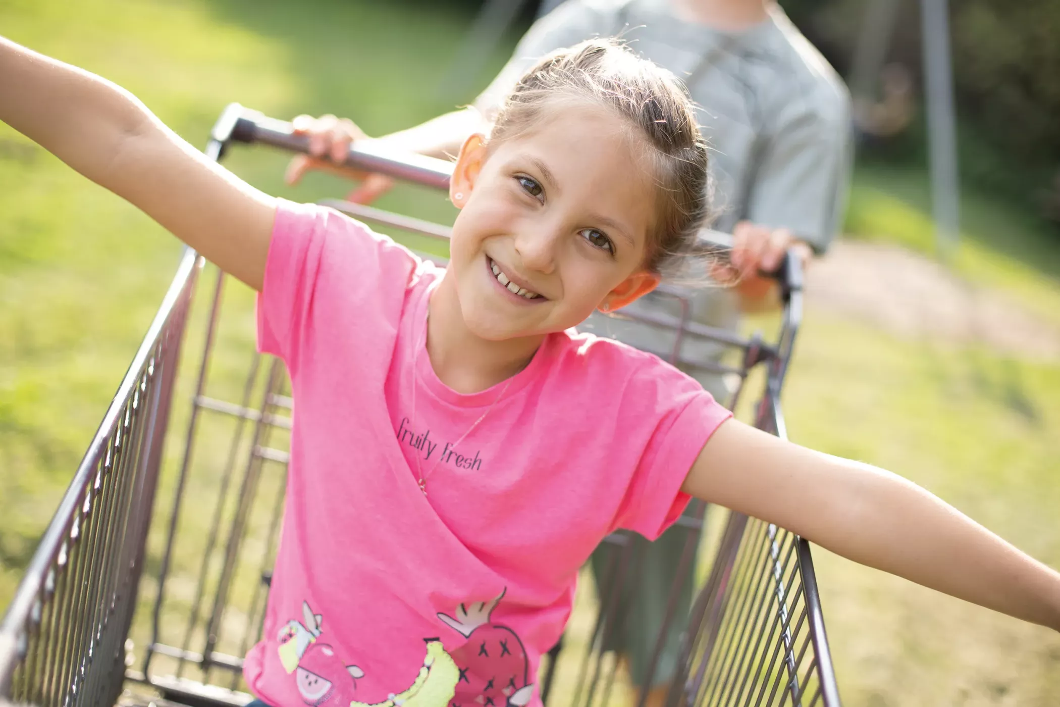 A smiling child in a pink shirt sits in a shopping cart in the open air while another child pushes the trolley behind him.