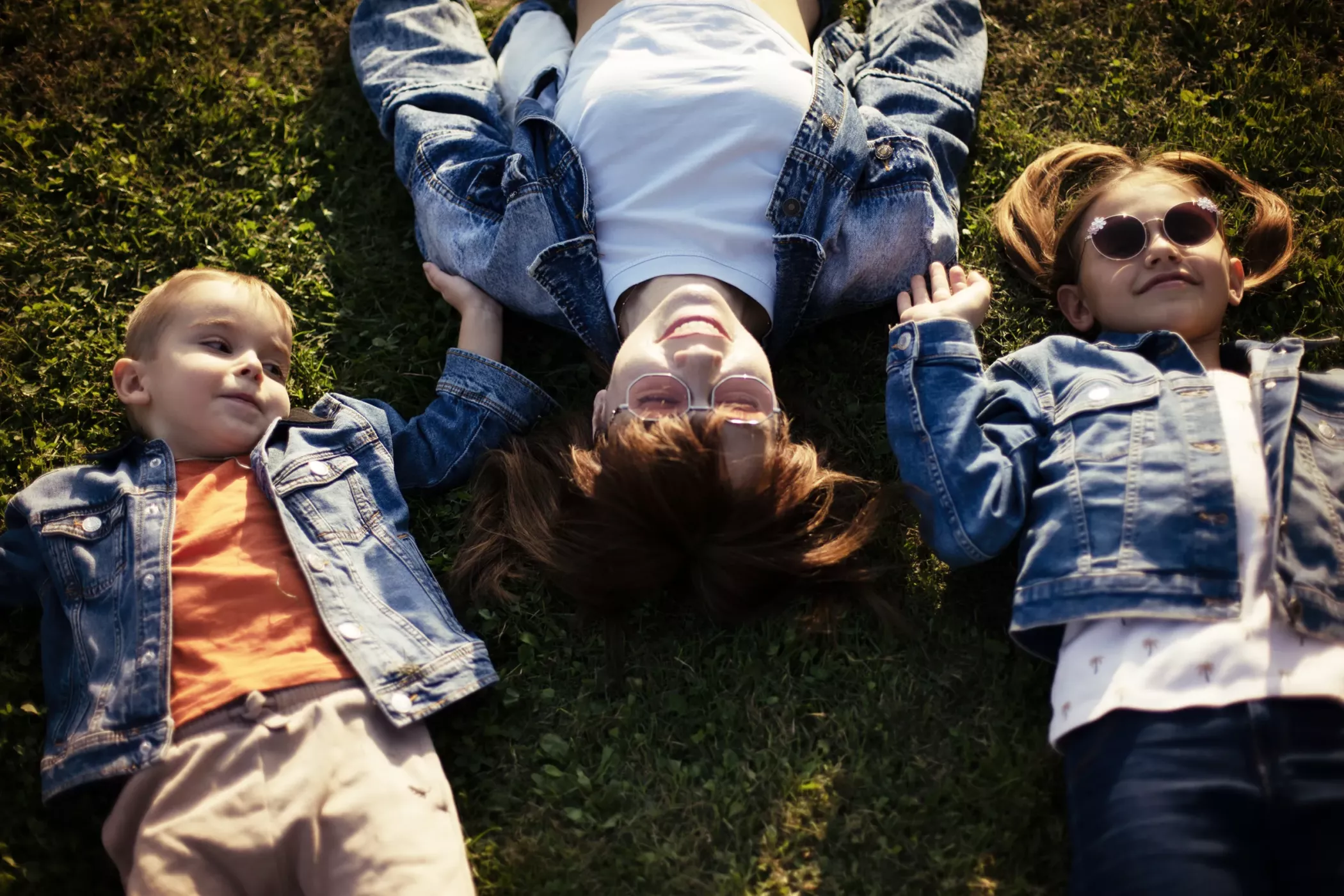 Three children lie on the grass, holding hands, smiling, wearing sunglasses and denim jackets in the sunshine.