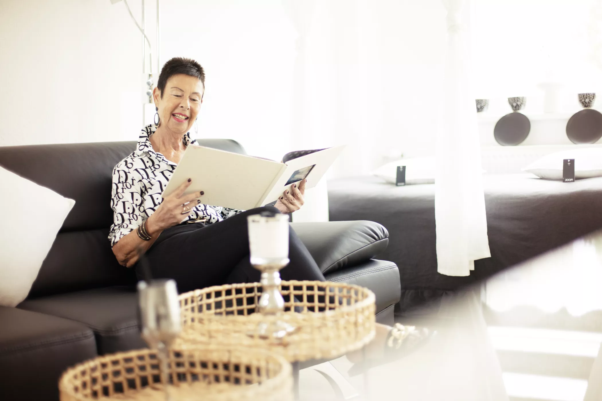 Eine Frau sitzt auf einem schwarzen Sofa in einem hellen Raum und liest lächelnd ein großes Buch. Bett und Tisch mit Kerzen im Hintergrund.