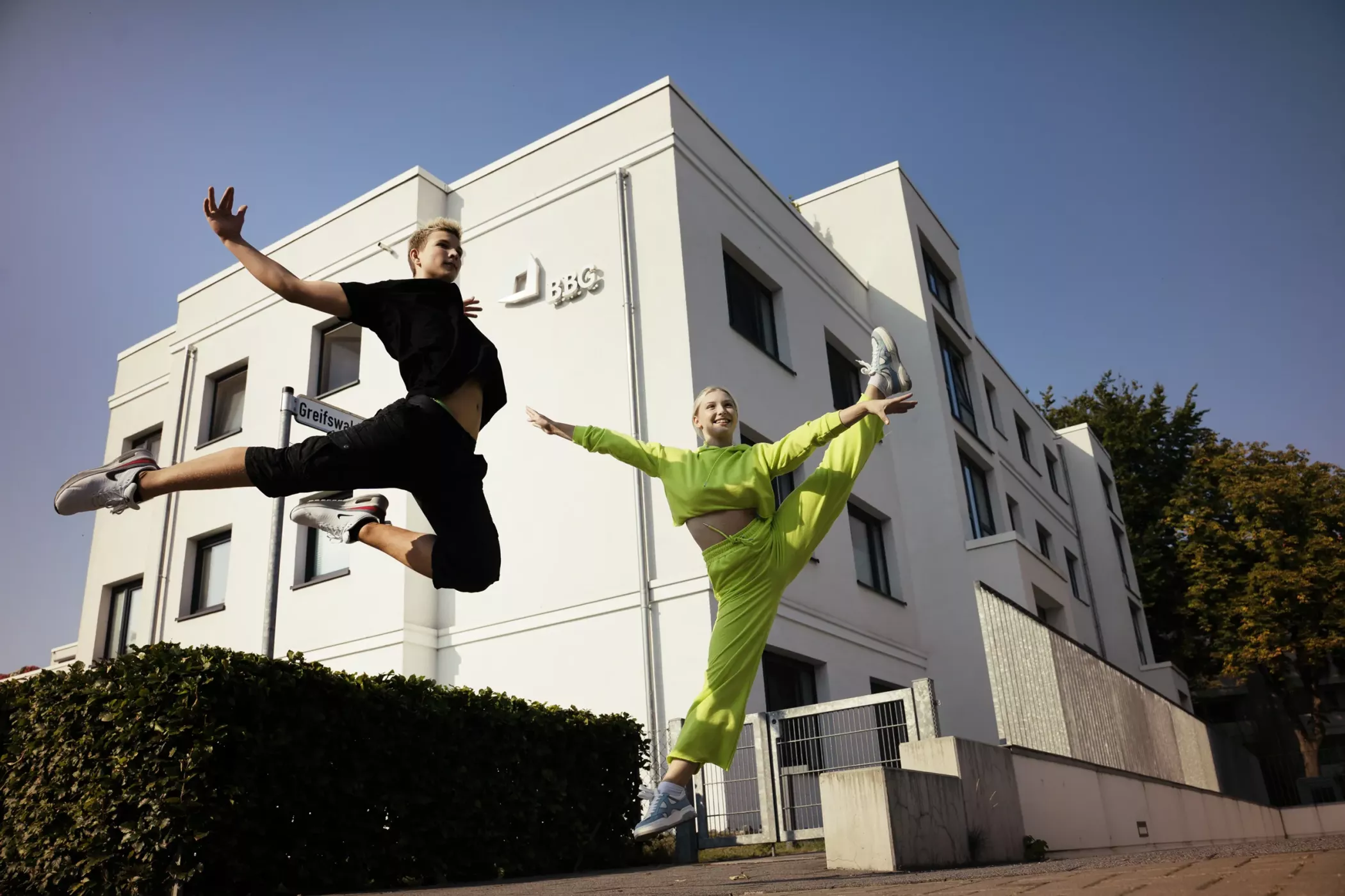 Zwei Personen in Sportkleidung vollführen an einem sonnigen Tag hohe Sprünge vor einem modernen weißen Gebäude.