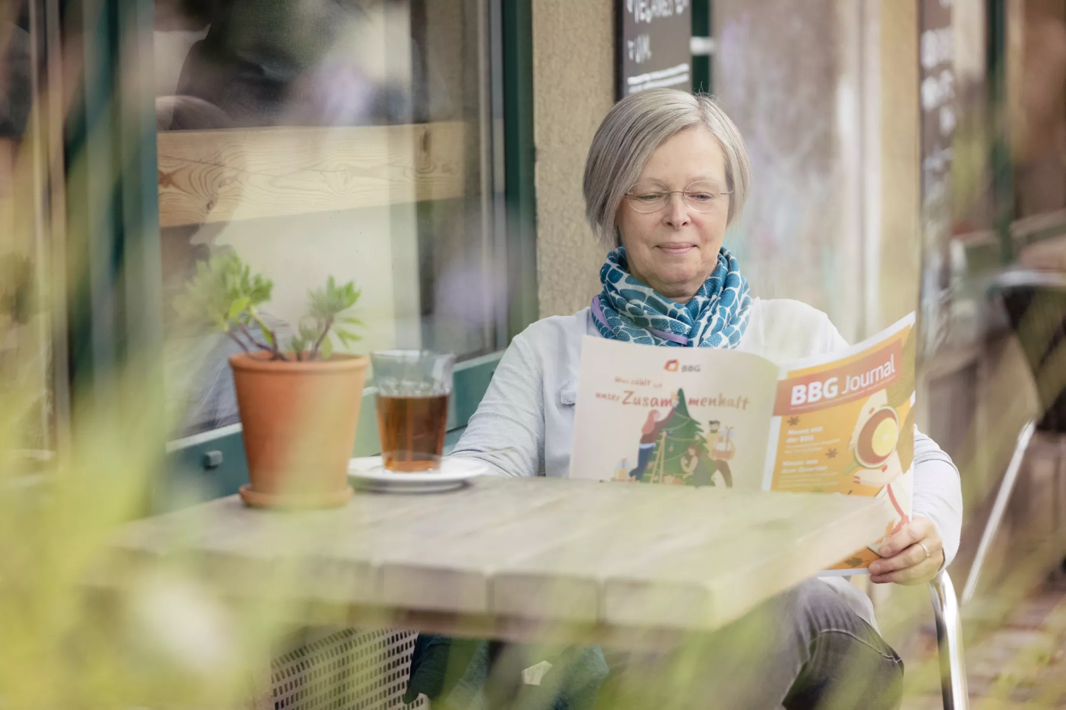 Eine alte Frau mit ergrauendem Haar liest an einem Cafétisch im Freien mit einer Topfpflanze und einer Tasse Tee eine Zeitschrift.