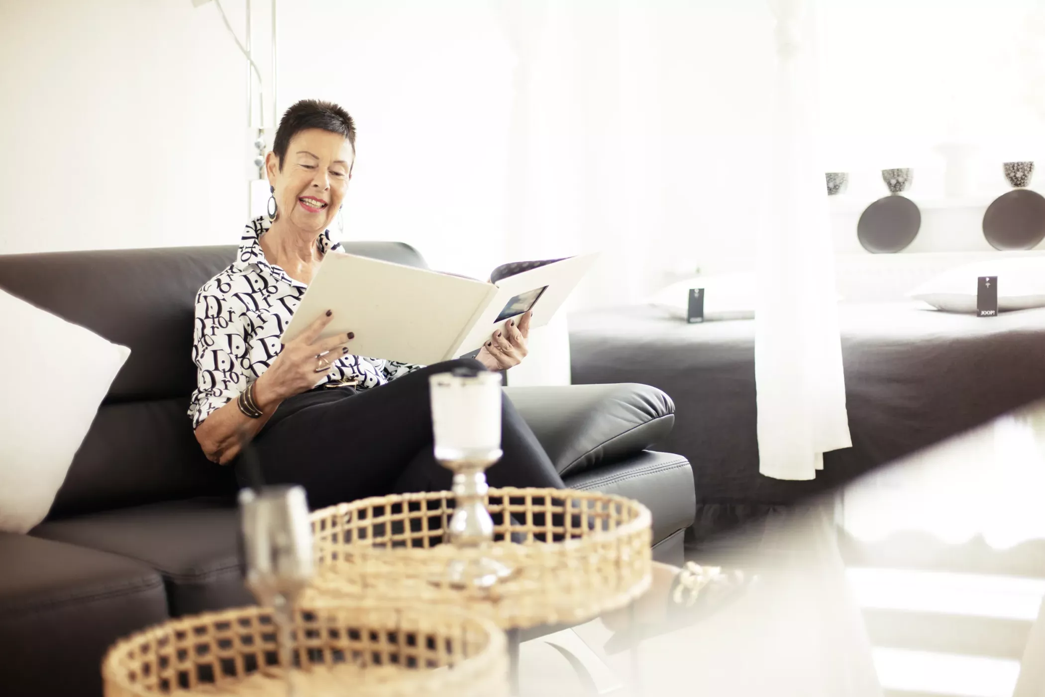 Açık renkler ve hasır masalarla dekore edilmiş aydınlık, modern bir oturma odasında gülümseyen bir kadın kanepede oturmuş kalın bir kitap okuyor.