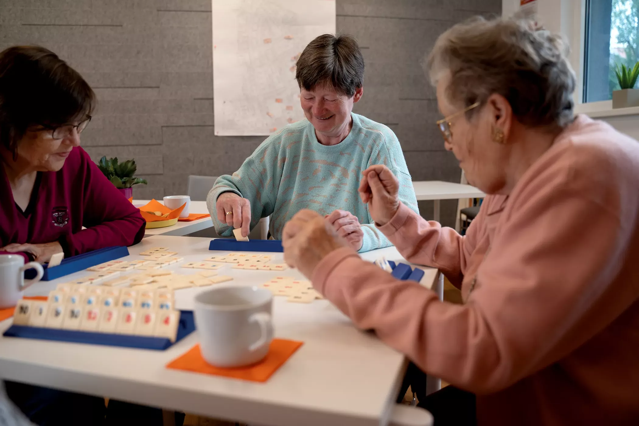 Drei ältere Frauen sitzen an einem Tisch und spielen zusammen ein Spiel, lächeln und genießen die Gesellschaft der anderen bei einem Kaffee.