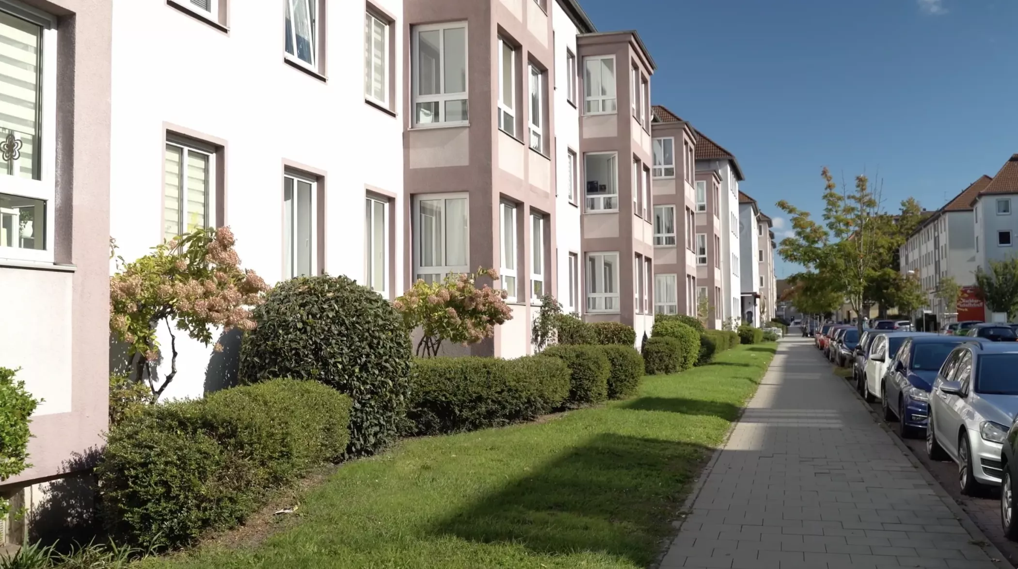 Eine Reihe moderner Wohnungen mit großen Fenstern, gepflegten Sträuchern und einem Bürgersteig an einem sonnigen Tag.