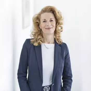 Une femme aux cheveux blonds bouclés, vêtue d'un blazer bleu marine et d'une chemise blanche, sourit dans un décor clair et simple.