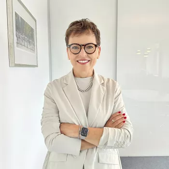 Lächelnde Frau mit kurzen Haaren und Brille, die eine helle Jacke und eine intelligente Uhr trägt, steht im Schneidersitz in einem hellen Büro.