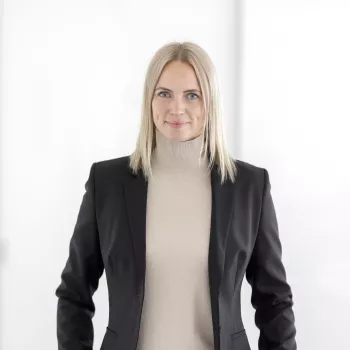 Une femme aux cheveux blonds et raides, portant un blazer beige et une veste noire, se tient devant un fond blanc uni et sourit légèrement.