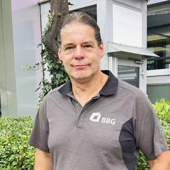 Ein Mann mit langen Haaren, der ein graues Polohemd der Marke BBG trägt, steht draußen in der Nähe von Sträuchern und einem modernen Gebäude.